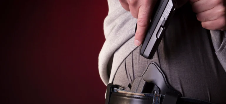 A person’s hand draws a black handgun from a holster attached to their waistband, with the background out of focus and red in color.