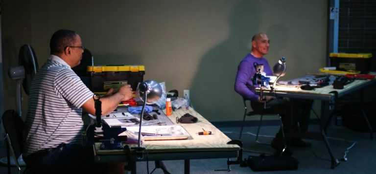 Two men sit at separate tables working on small mechanical parts under desk lamps in a dimly lit room, tools and cases spread out before them. One man is smiling, perhaps proud after earning his POST AR armorer certification in St. Louis.