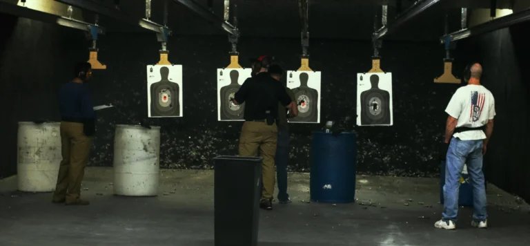 Three people stand in an indoor shooting range during a basic shotgun class in St. Louis, facing paper target silhouettes. Two barrels and a trash bin are nearby, while spent bullet casings litter the floor.