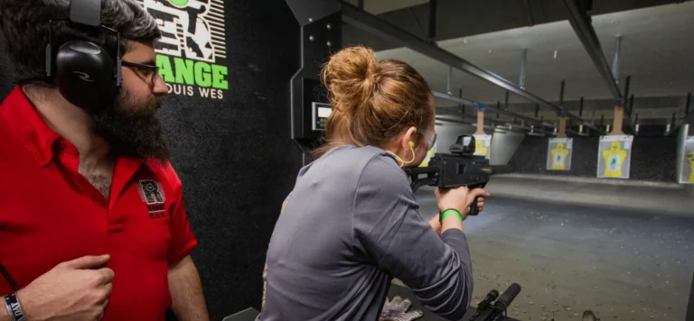 A woman participates in rifle carbine training at an indoor shooting range in St. Louis, aiming at targets while a man in a red shirt and ear protection observes beside her. Targets are visible down the range.