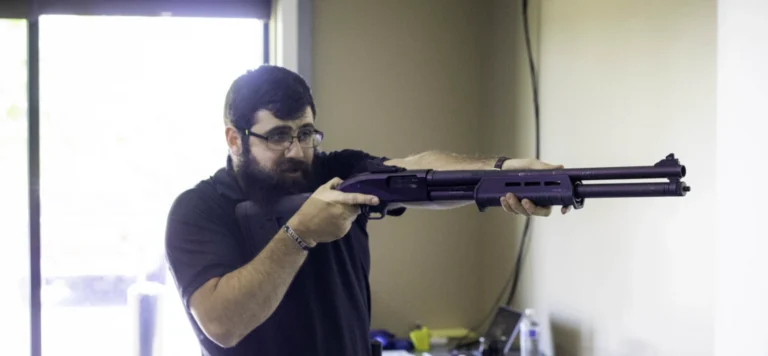 A man with a beard and glasses aims a shotgun indoors, standing near a window with light coming through. Wearing a black shirt, he appears focused—possibly practicing for a shotgun training class in St. Louis.