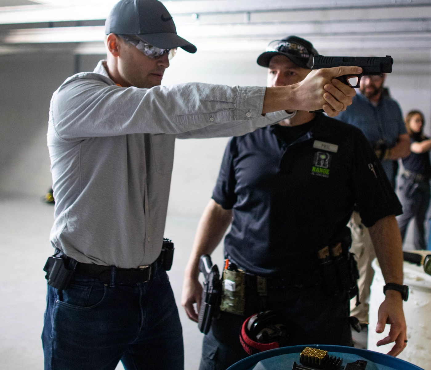 A man wearing safety glasses aims a handgun at an indoor shooting range during an intermediate pistol course St. Louis, while an instructor in a black shirt observes closely. Another person stands in the background with ammunition visible on a nearby barrel.