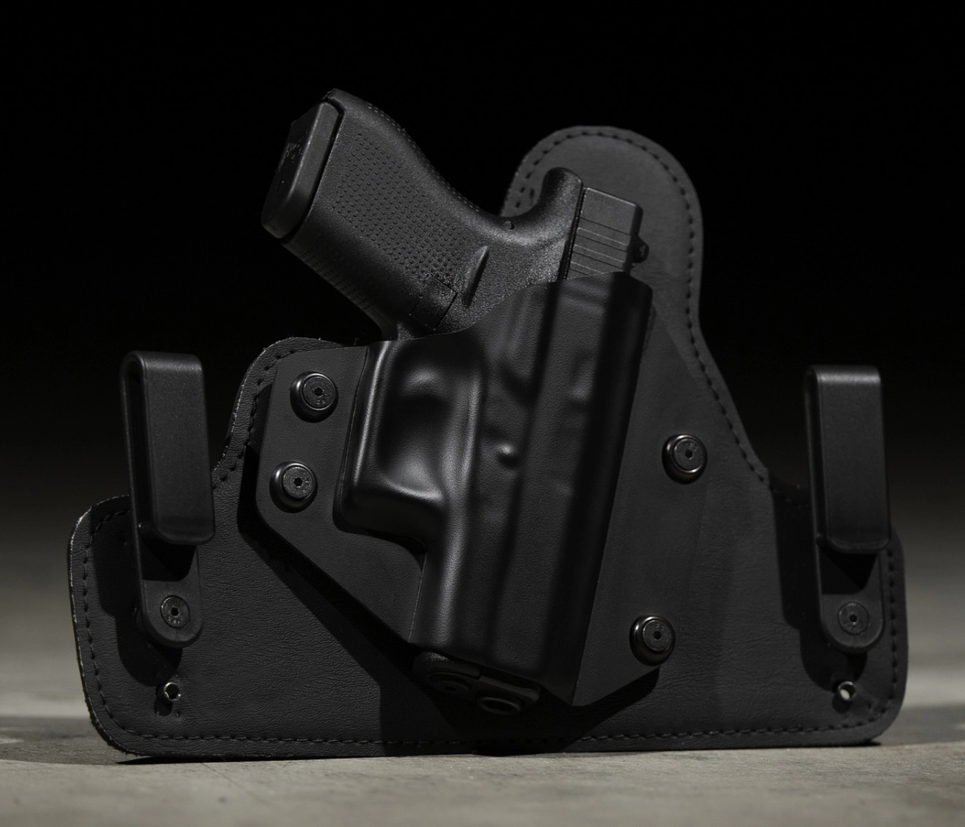 A black handgun in a black holster with dual clips, resting on a dark surface against a black background.