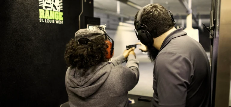 Two people wearing ear protection stand at an indoor shooting range during a basic handgun class St. Louis. One person aims a handgun at a target while the other, possibly an instructor, observes and provides guidance. “Range St. Louis West” is seen on a sign.