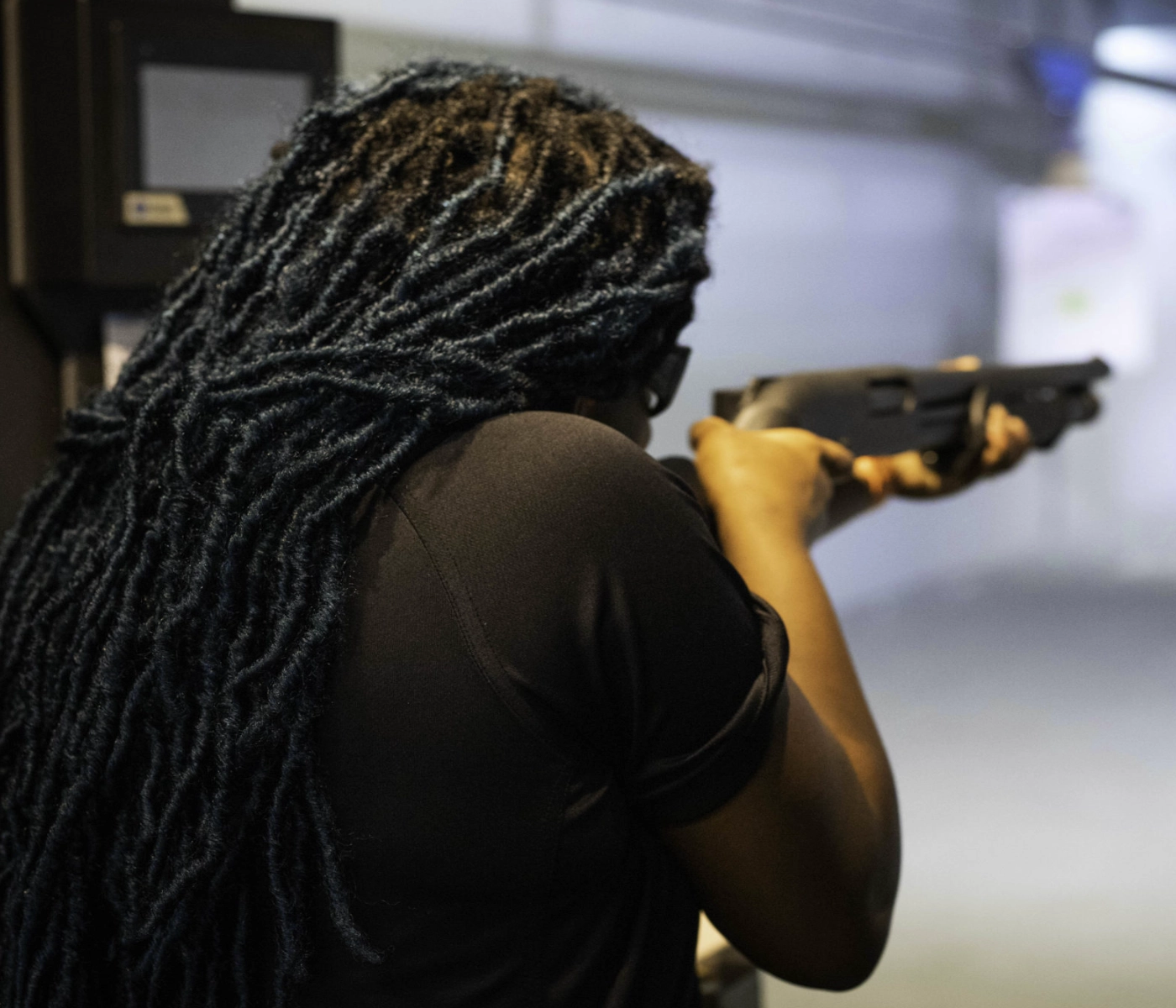 A person with long, dark, tightly twisted hair wearing a black shirt aims a shotgun at an indoor shooting range during a basic shotgun class in St. Louis, facing away from the camera.