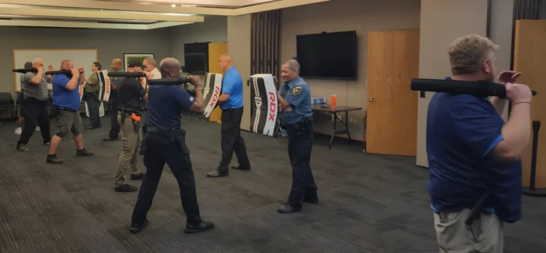A group of people, including police officers, participate in a POST baton certification St. Louis self-defense training session indoors, holding padded batons and using shields for practice. The room has dark carpet, TVs, and wooden doors.