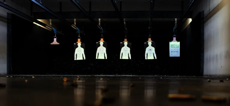 A dimly lit indoor shooting range, set up for an advanced rifle carbine course in St. Louis, features five illuminated human-shaped paper targets and a grid-style target, viewed from a low angle with scattered shell casings on the floor.