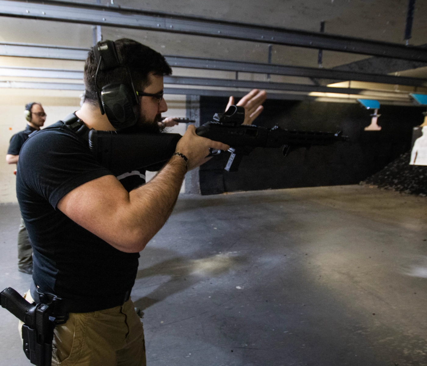 A man wearing ear protection aims a rifle at an indoor shooting range during an advanced rifle carbine course in St. Louis, with another person in the background and targets visible at the far end.