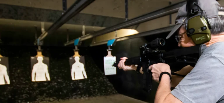 A person wearing ear protection aims a rifle at human-shaped paper targets during a basic rifle course St. Louis at an indoor shooting range.