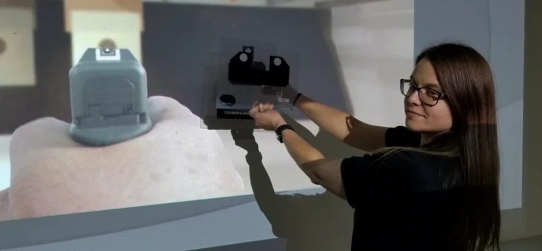 A woman wearing glasses points a gun-shaped training device at a projection of a handgun aiming at a target on a wall, participating in a simulated firearms training exercise for CCW certification Missouri.