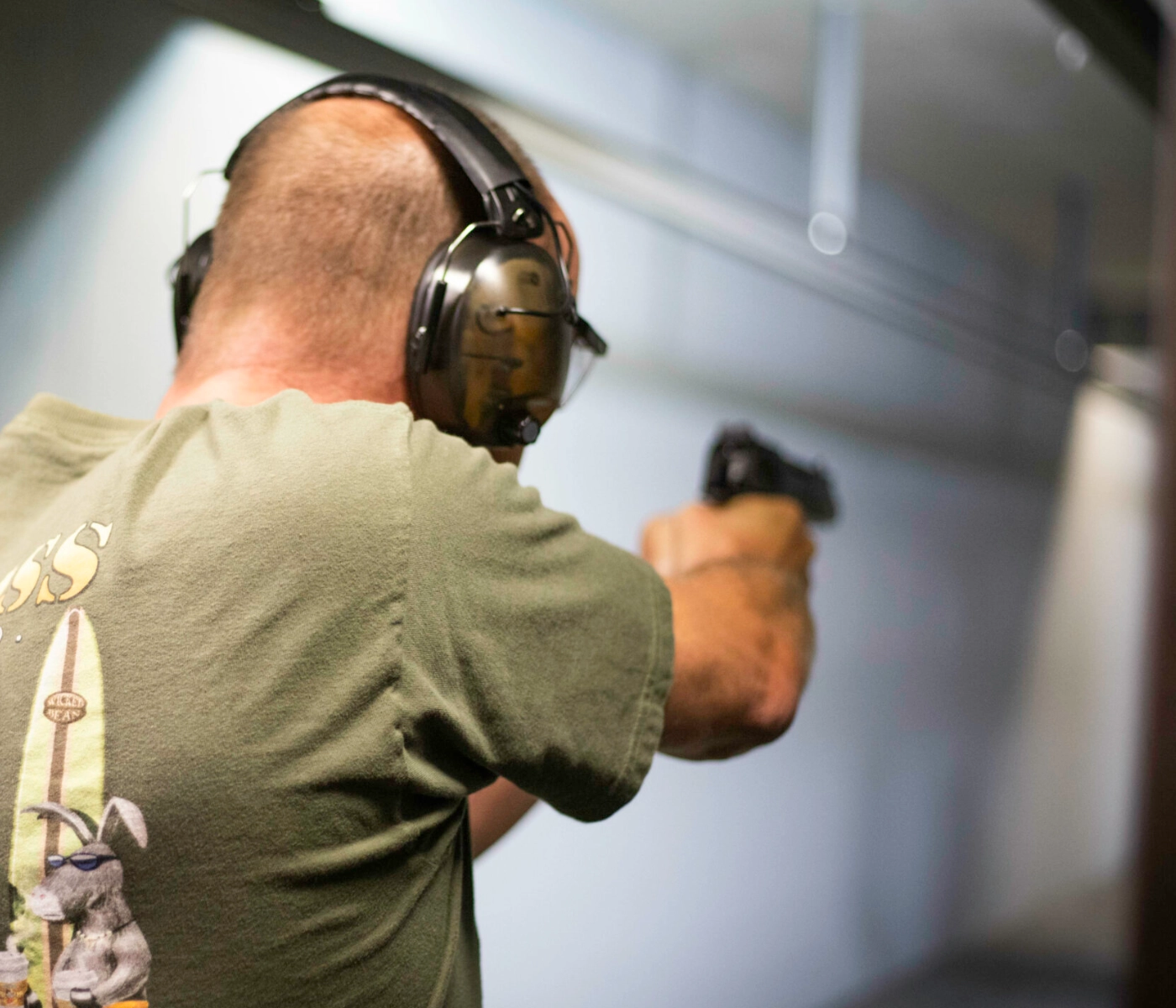 A person wearing ear protection and a green T-shirt aims a handgun at an indoor shooting range, possibly practicing for their CCW certification in Missouri, with their back to the camera.