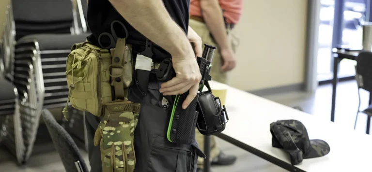 A person in tactical gear, possibly attending an advanced defensive handgun course St. Louis, places a handgun into a holster on their hip. They have scissors, pouches, and camouflage accessories on their belt, while another stands in the background near a table and chairs.