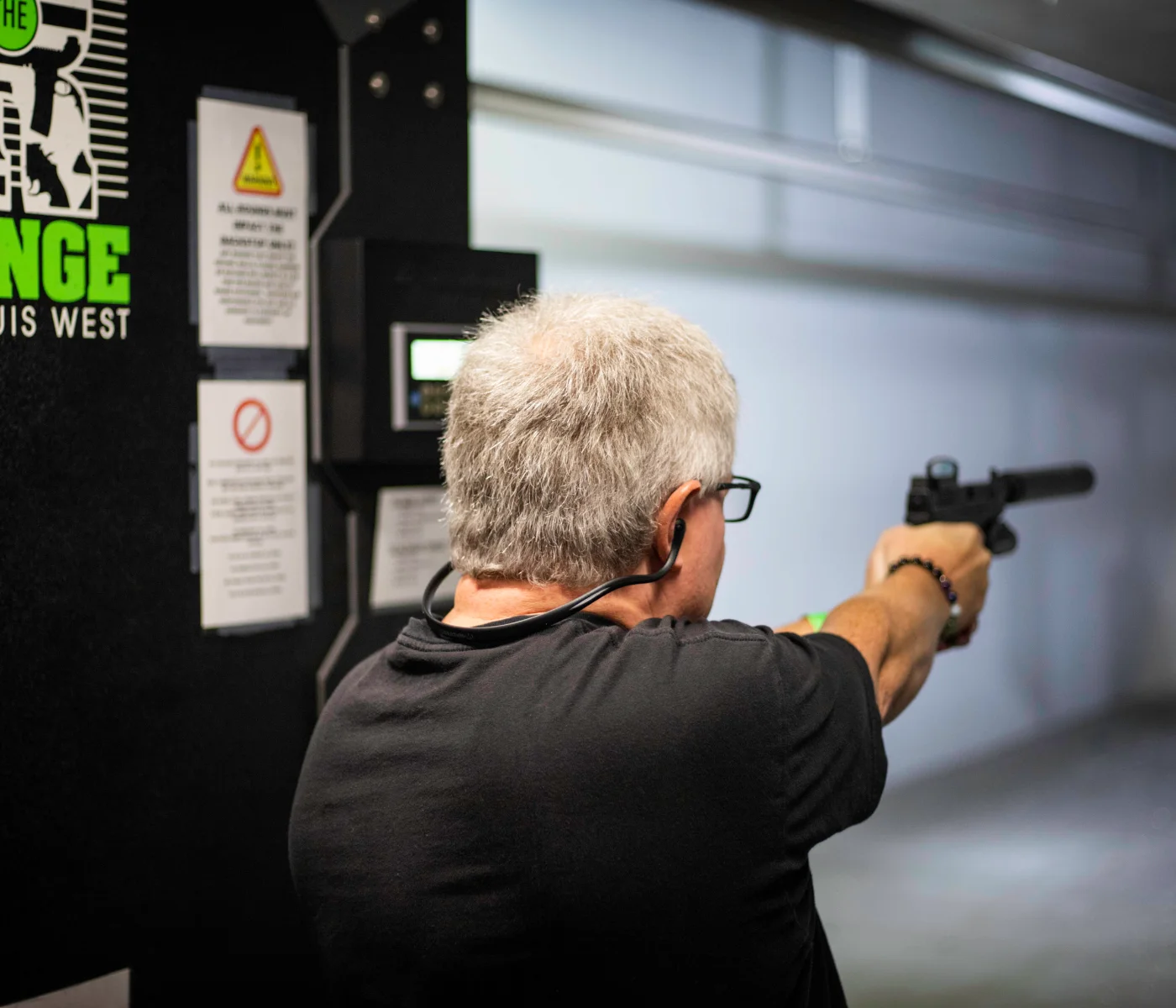 A man with gray hair and glasses aims a handgun at an indoor shooting range during an advanced defensive handgun course in St. Louis, wearing protective ear gear and standing near safety instructions posted on the wall.