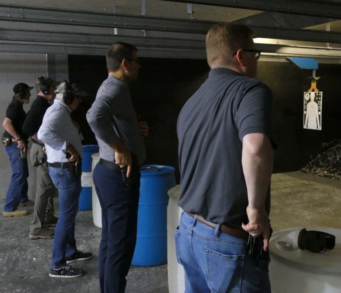 Four men stand indoors at a shooting range, each with a handgun holstered at their waist, facing a human silhouette target. Blue barrels and tables are nearby, and they appear to be preparing to shoot.