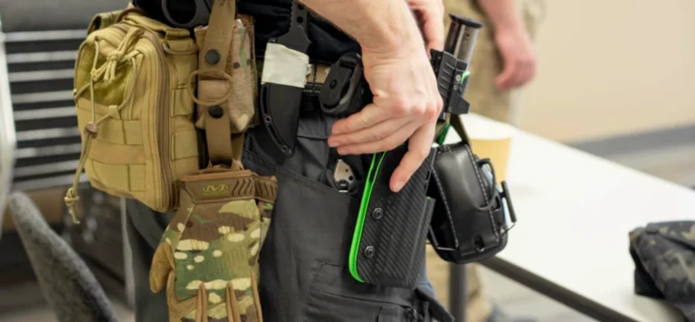 A close-up of a person wearing tactical gear, including a holster, pouch, gloves, and utility belt, as they adjust or draw a handgun from the holster.