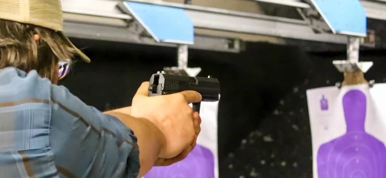 A person wearing a cap aims a handgun at a purple human-shaped target during a defensive handgun level 2 session at an indoor St. Louis shooting range. The focus is on the shooter’s arms and gun, with the target blurred in the background.