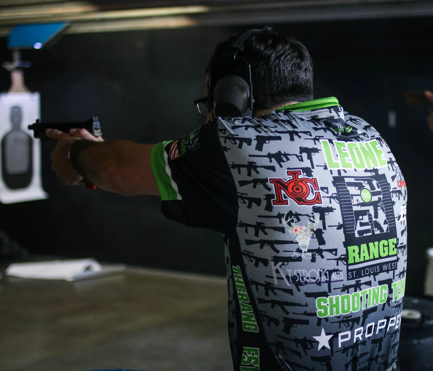 A person in a graphic shooting team jersey and hearing protection aims a handgun at a paper target during a defensive handgun course St. Louis at an indoor range. The focus is on the shooter from behind.