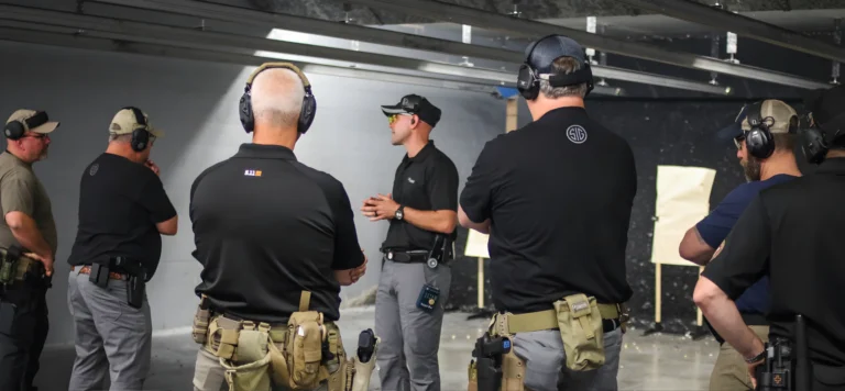 A firearms instructor speaks to a group of people wearing ear protection and tactical gear at an indoor shooting range, where OC spray certification St. Louis courses are also offered, with paper targets visible in the background.