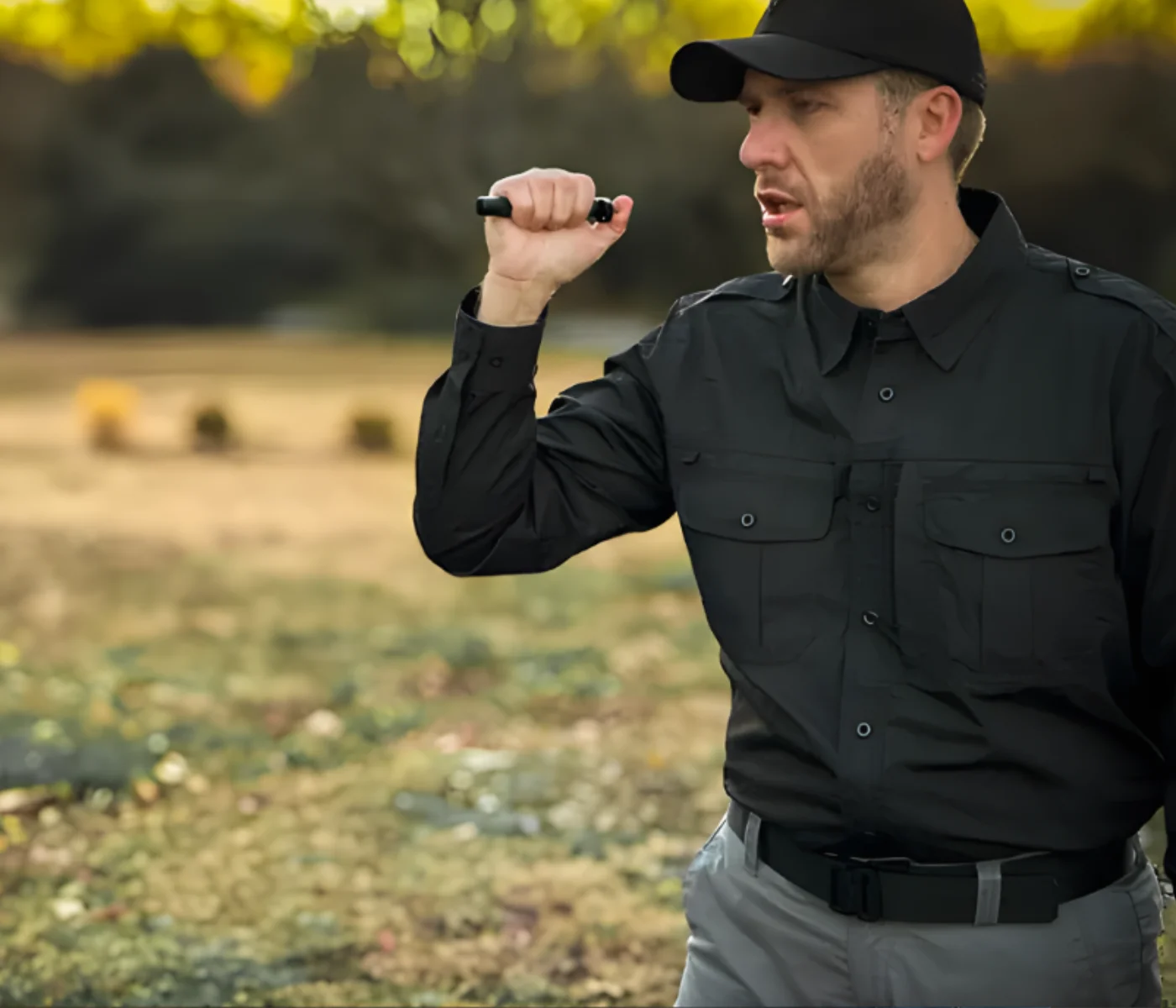 A man in a dark uniform and cap stands outdoors, holding a walkie-talkie near his mouth, appearing to communicate—possibly during OC spray certification training in St. Louis. The background is blurred greenery and sunlight.