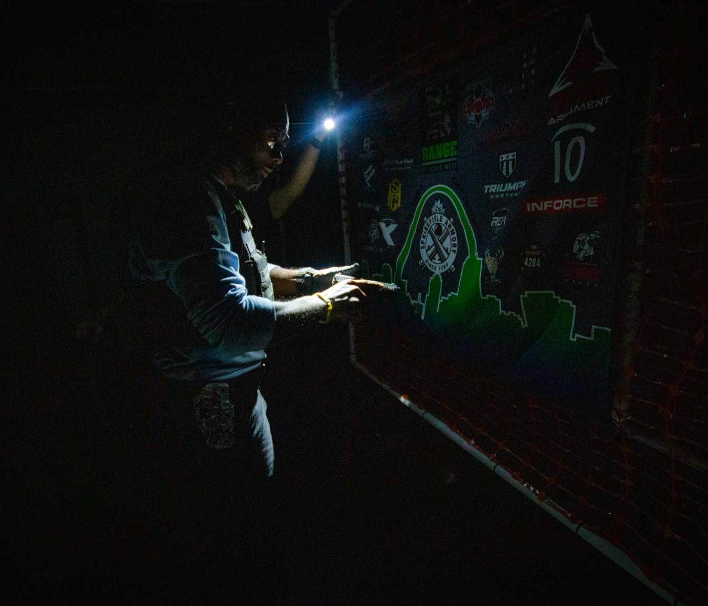 A person uses a headlamp to illuminate and point at a colorful board with various logos in a dark environment, suggesting careful inspection or navigation—much like the focused training in a low light shooting course St. Louis offers.
