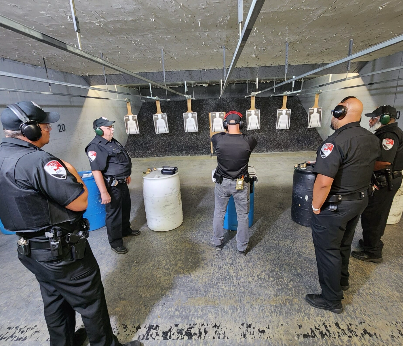 Five security officers in uniforms, ear protection, and holsters stand at an indoor shooting range for a practical pistol course in St. Louis. One officer aims at a target while the others observe.
