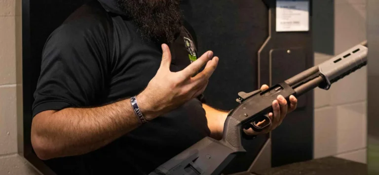 A person with a dark beard wearing a black shirt holds a shotgun and gestures with their other hand, possibly explaining or demonstrating POST shotgun qualification indoors in St. Louis.