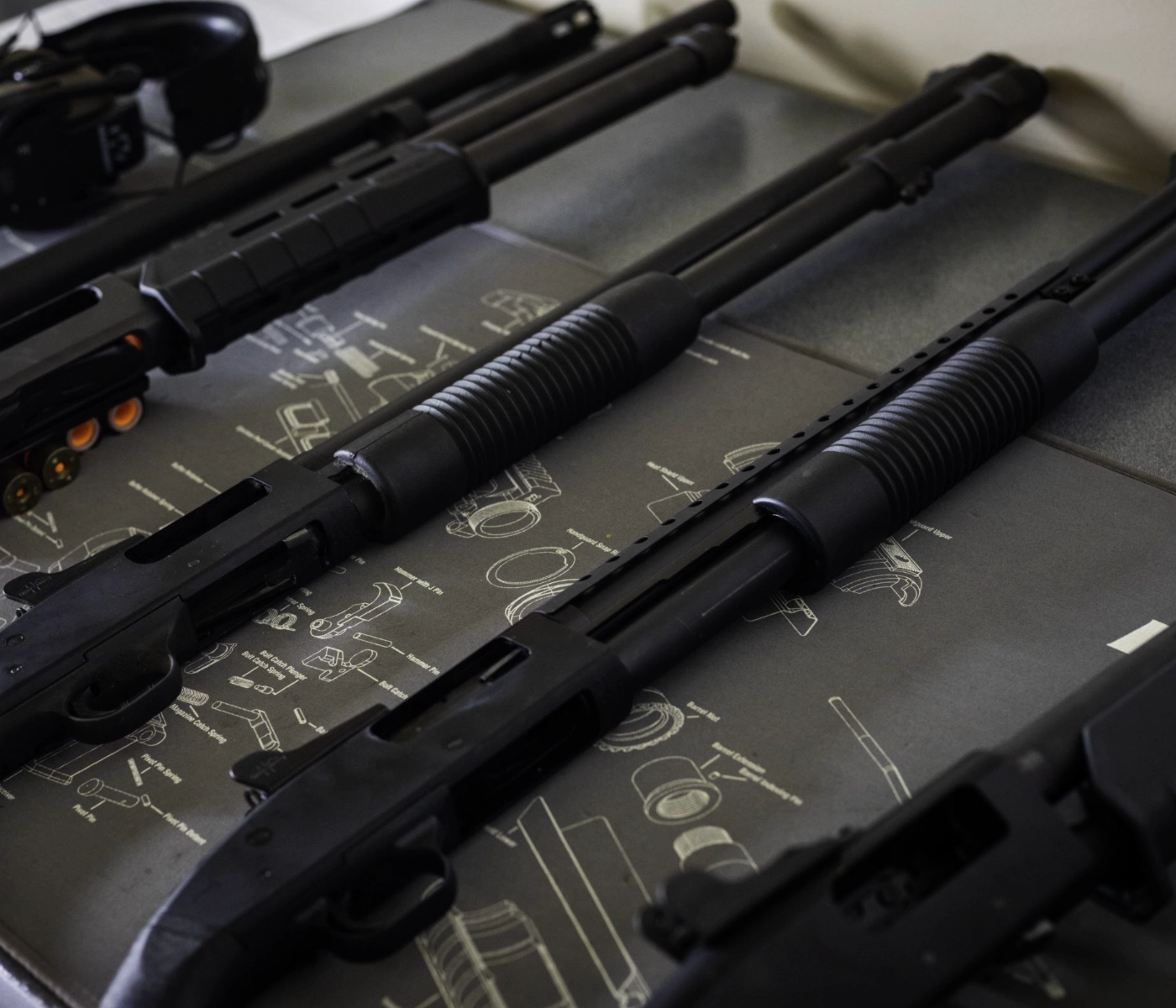 Three black pump-action shotguns are laid out side by side on a gun cleaning mat with assembly diagrams, ready for a shotgun training class St. Louis participants will appreciate. Parts of other guns and accessories are visible around the shotguns.