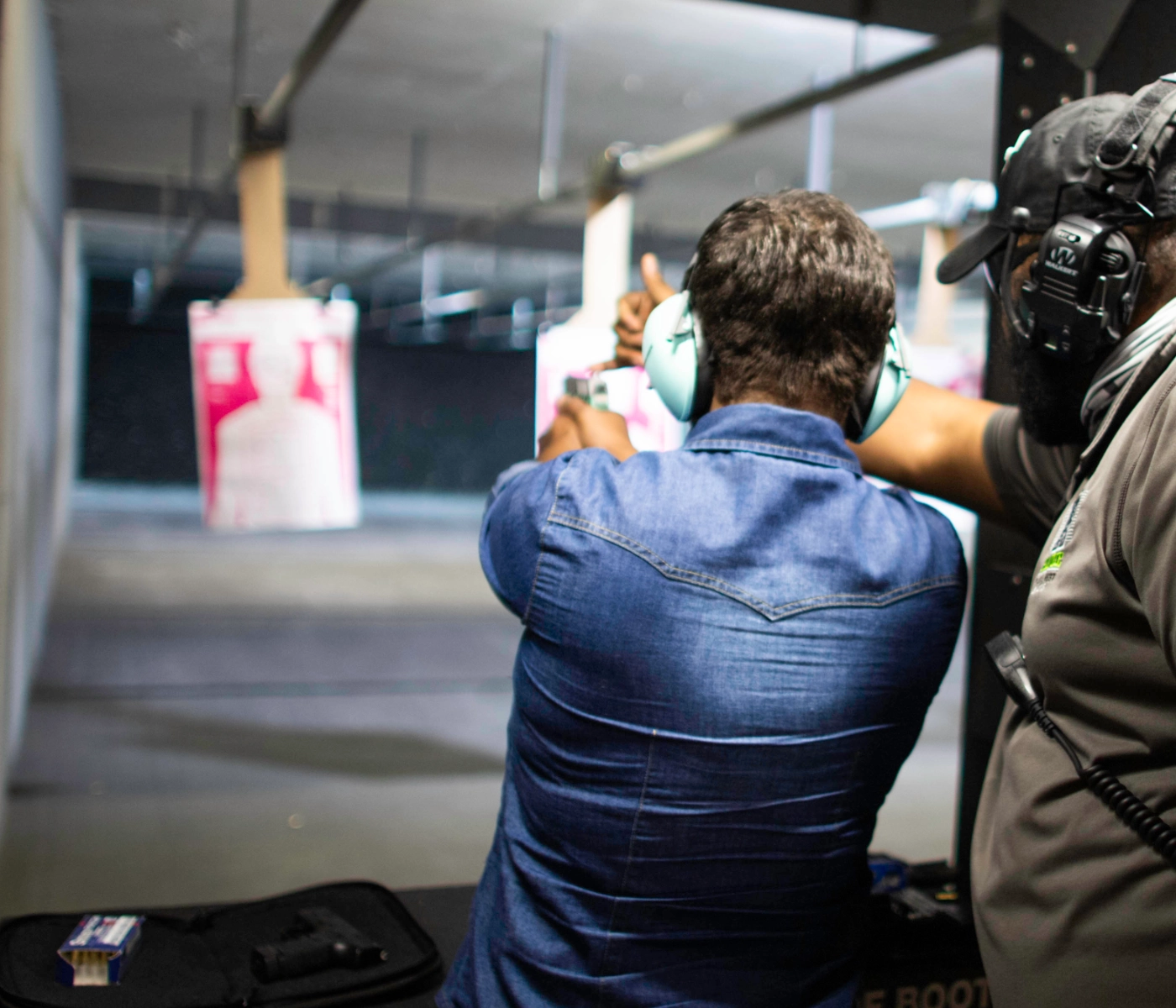 Two people at an indoor shooting range; one is wearing headphones and aiming a handgun with red dot sight training, while the other, also with headphones, stands beside and instructs him in St. Louis.