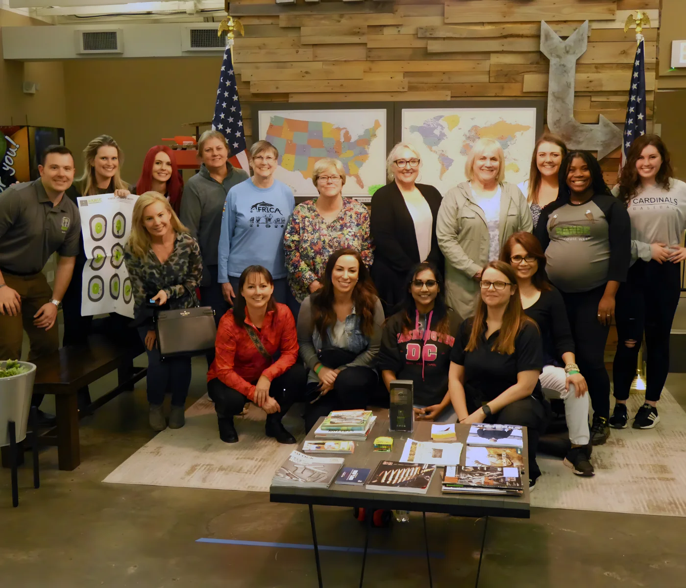 A group of sixteen people, mostly women, pose and smile together indoors in front of two U.S. flags and a wall with maps. Some are standing, others kneeling, and various items and pamphlets are on a table in front of them.