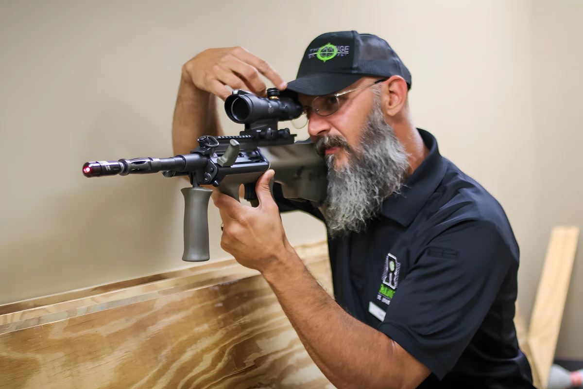 A bearded man wearing glasses, a black cap, and a black shirt aims a rifle with a scope indoors, resting the rifle on a wooden ledge while adjusting the scope.