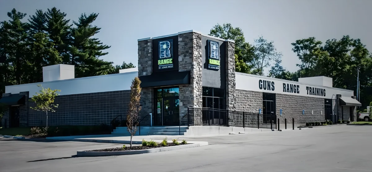 A modern, single-story building with signs reading "Range," "Guns," and "Training" on the exterior. The entrance is framed by stone accents and the parking lot is mostly empty. Trees are visible in the background.
