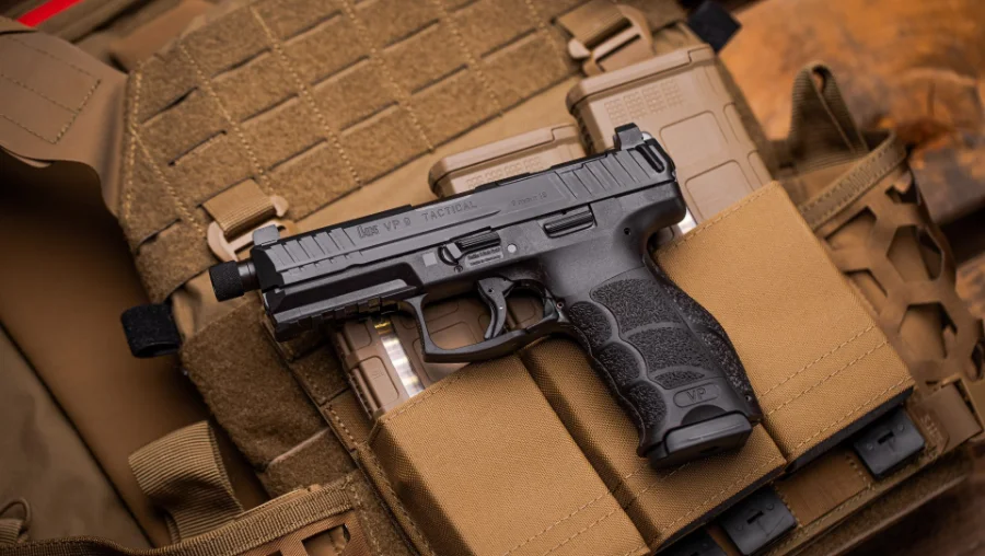 A black semi-automatic handgun rests on top of a tan tactical vest with magazine pouches, surrounded by additional magazines and gear.