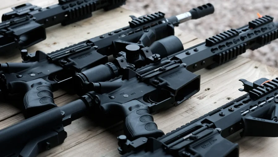 Several black semi-automatic rifles are arranged side by side on a wooden table, displaying their barrels, handguards, scopes, and grips in close detail.