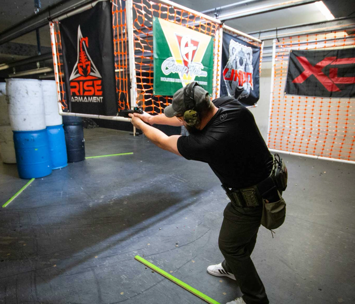 A person in a black shirt and cap aims a handgun at an indoor shooting range during advanced pistol drills St. Louis, positioned behind orange netting and surrounded by barrels and banners promoting firearm brands.