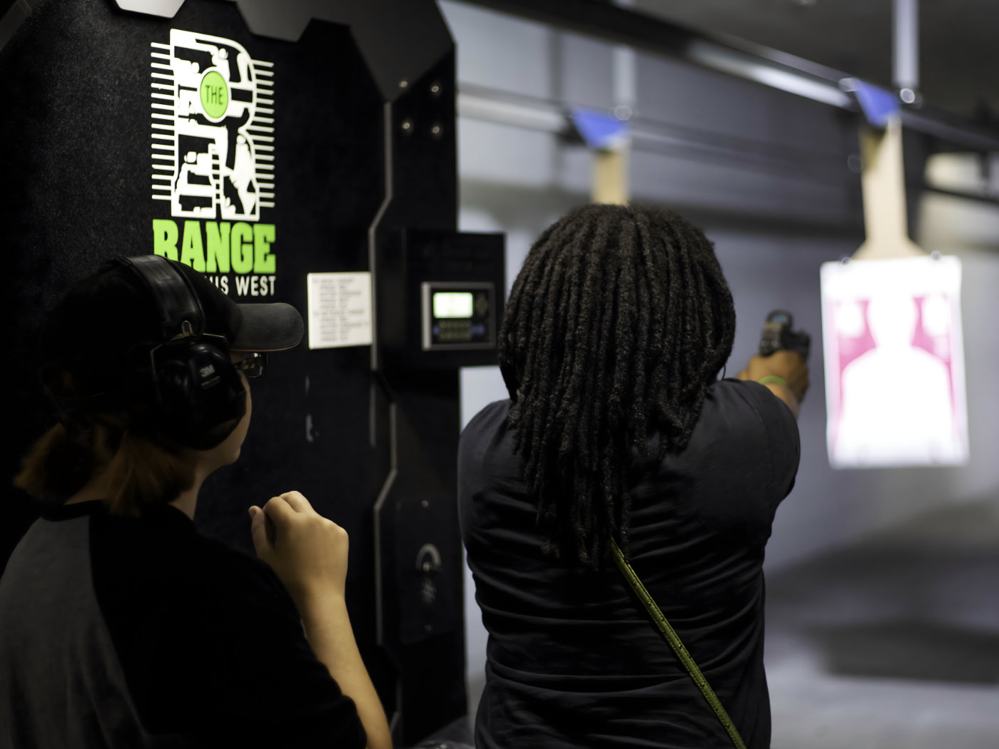 Two people at an indoor shooting range, one aiming a handgun at a paper target while the other watches. Both wear hearing protection. The range sign and electronic equipment are visible on the wall.