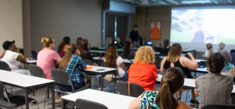 A group of people sit at desks in a classroom, facing a projected screen at the front where a person presents during a women's self defense seminar in St. Louis. Some attendees take notes while others watch the screen with interest.